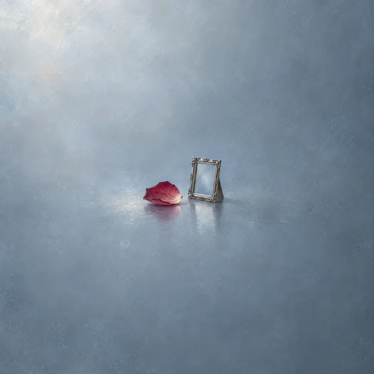 A pressed rose petal on frosted glass beside a small empty silver frame.