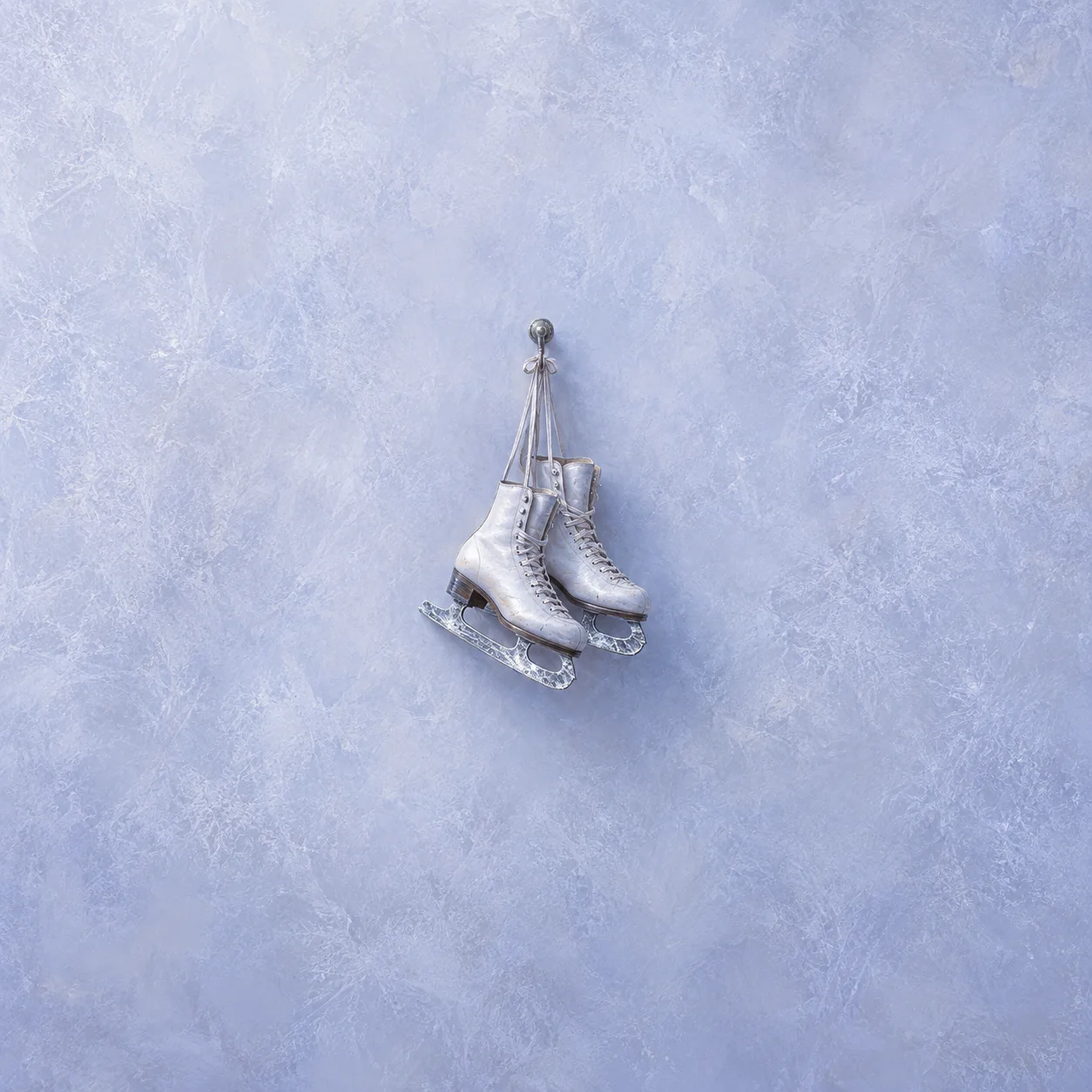 A pair of white ice skates hangs by its laces against a scratched pale lilac surface.
