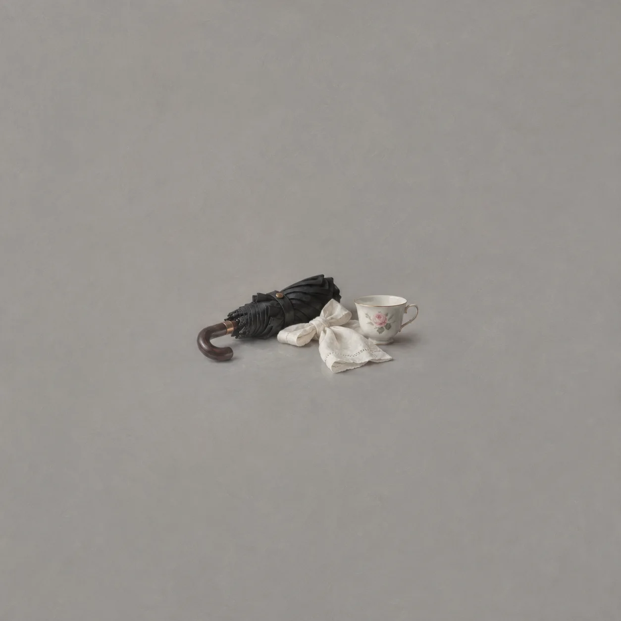 A folded black umbrella lies beside a floral teacup and a tied white napkin against grey.