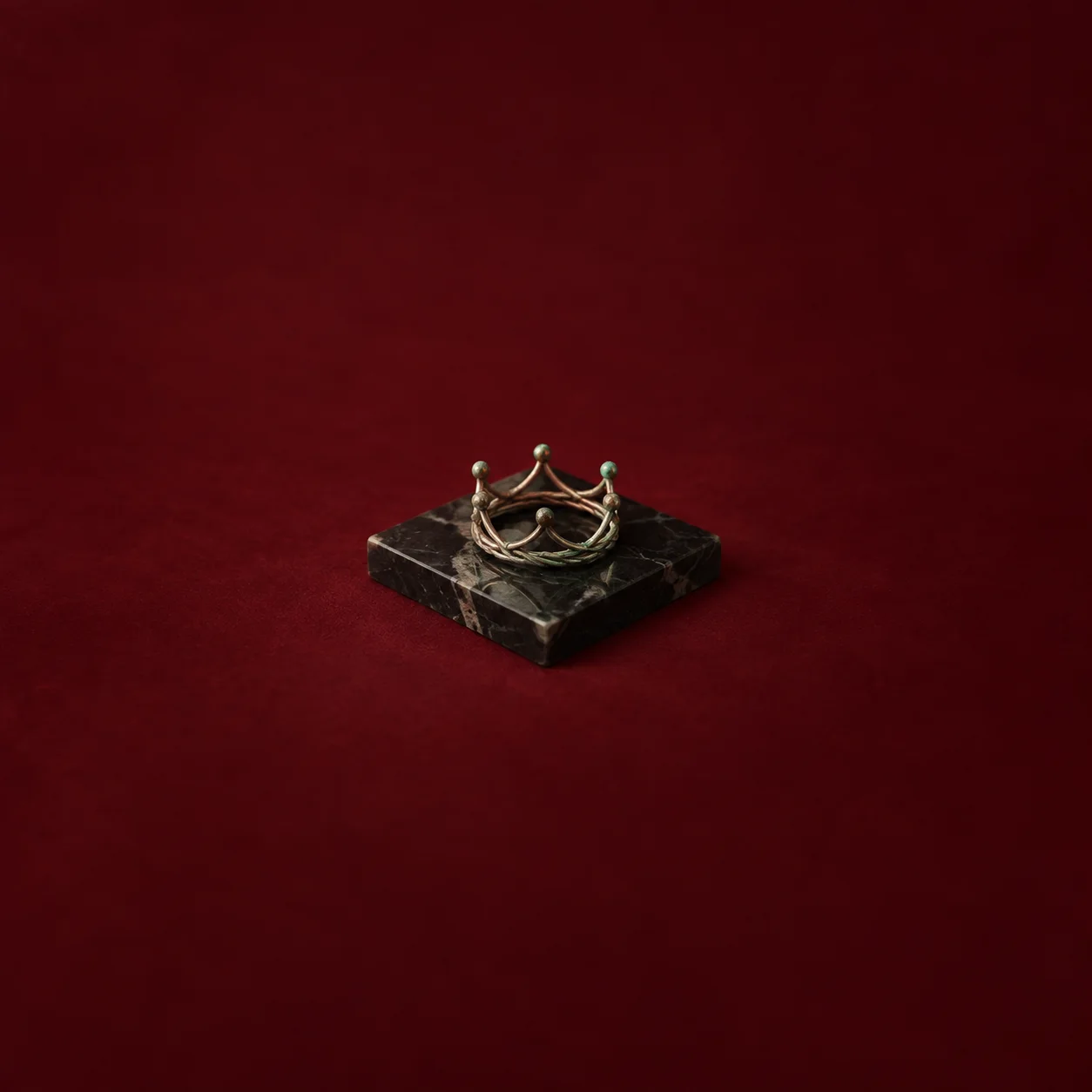 A small tarnished crown-shaped metal ring rests on a dark marbled slab against a burgundy background.