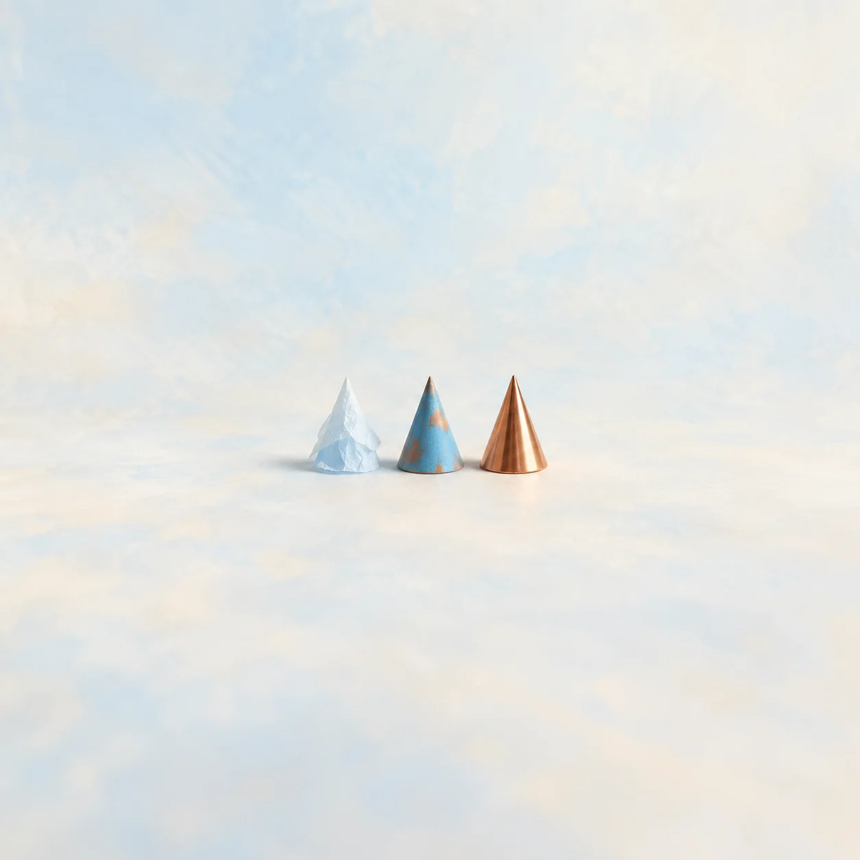 Three small cones in a row, made of tissue paper, cardboard and polished copper, against a pale sky-blue background.