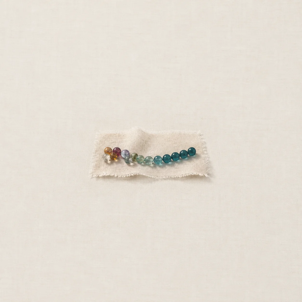 A curved line of glass beads shifts from mixed colours to teal on a strip of linen.