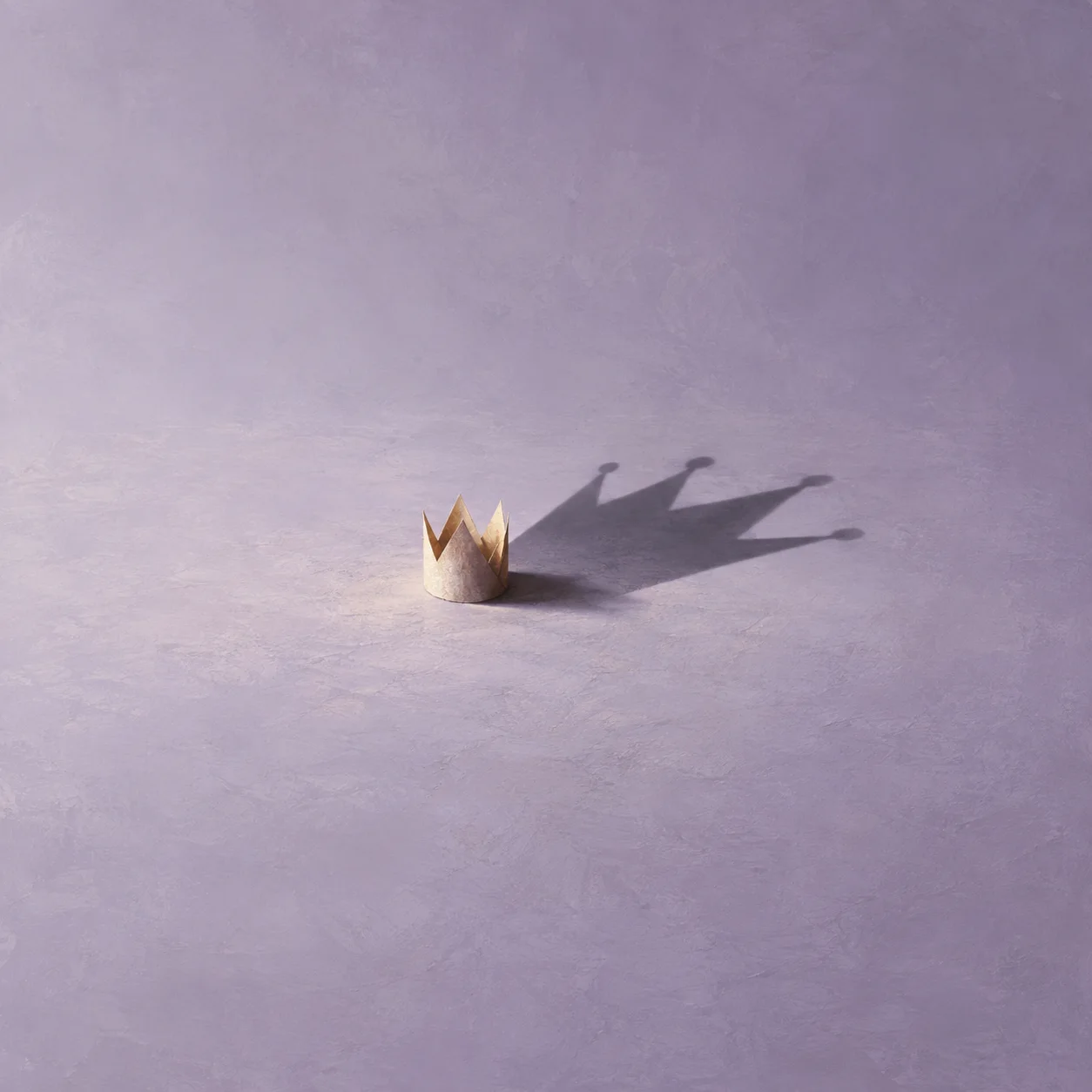 A small paper crown casting a much larger crown-shaped shadow across a lavender surface.