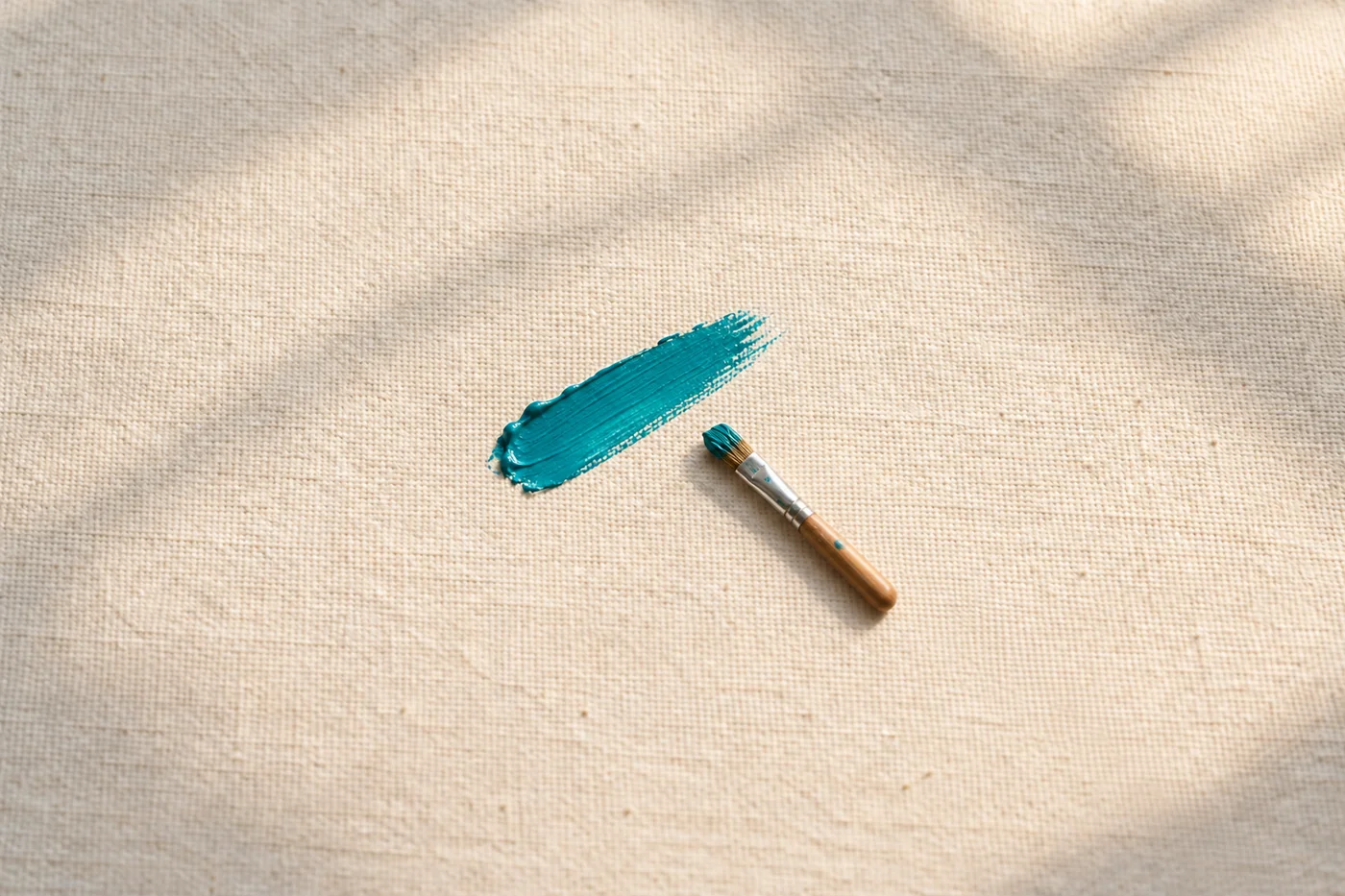 A short paintbrush lies beside one teal brushstroke on cream canvas.