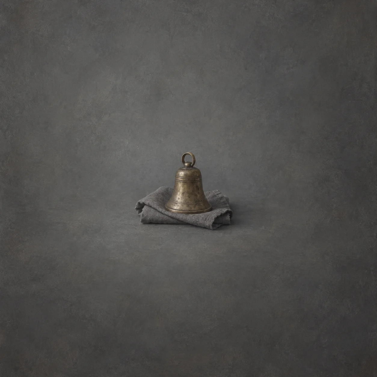 A tarnished brass bell rests on folded grey cloth against charcoal.