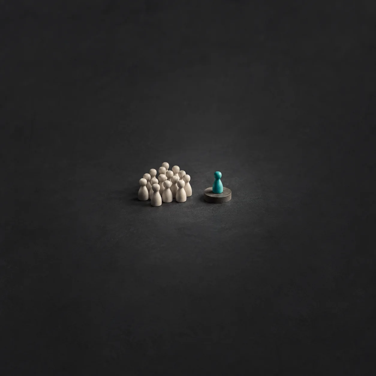 A group of small white wooden figures with one teal figure standing apart on a round platform against a dark background.