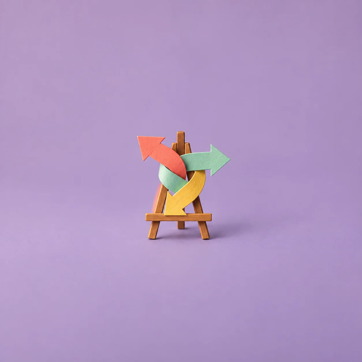 Coral, mint and yellow paper arrows overlap in different directions on a small wooden easel against lilac.