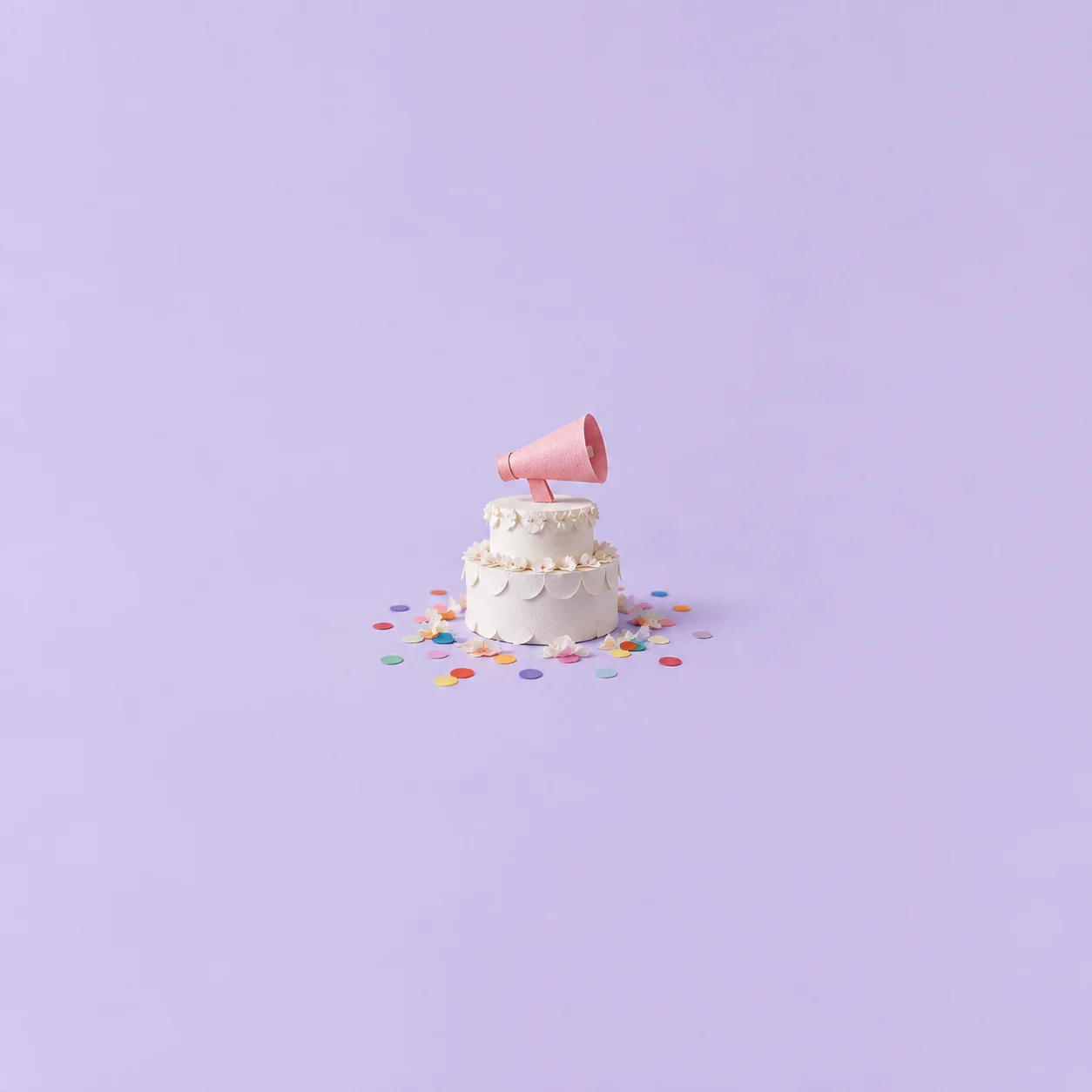 A small white tiered cake topped with a pink megaphone stands amid colourful confetti on lavender.