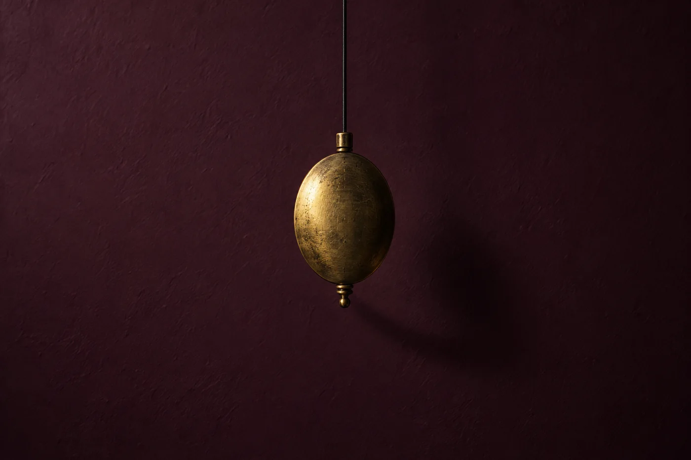 An oval brass pendulum hangs against a plum wall, lit along one edge.