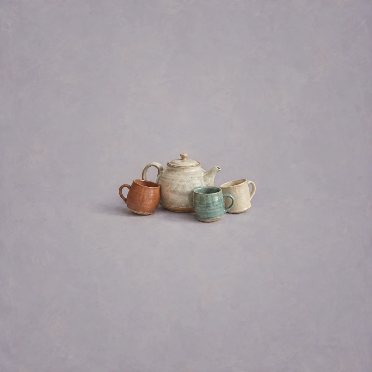 Three glazed mugs gathered around a teapot on a grey-lilac background.