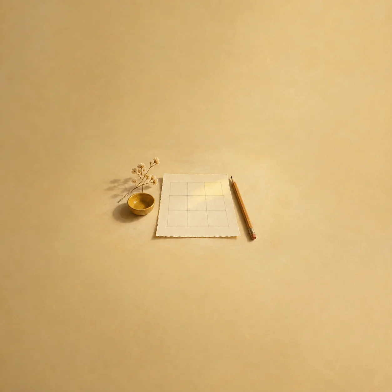A pencil lies beside a blank grid on a sheet of paper with a small gold bowl and dried sprig on a mustard-yellow surface.