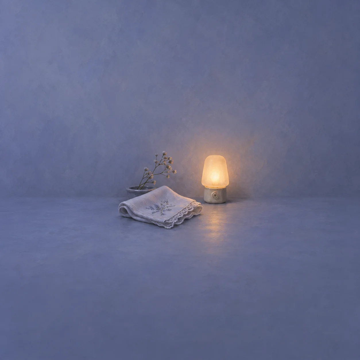 A folded pale handkerchief rests beside a glowing amber nightlight and a small dried sprig on periwinkle ground.