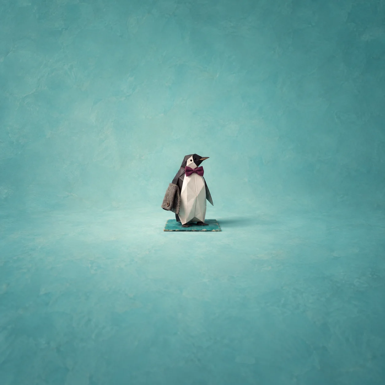 A folded paper penguin wearing a plum bow tie with a tweed patch at one wing, standing on a turquoise square.