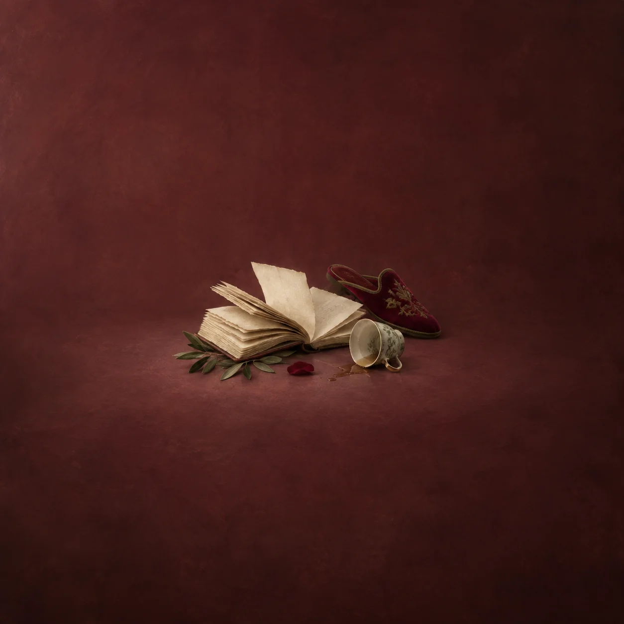 Loose pages fan out beside a velvet slipper, a tipped teacup and scattered leaves on burgundy.