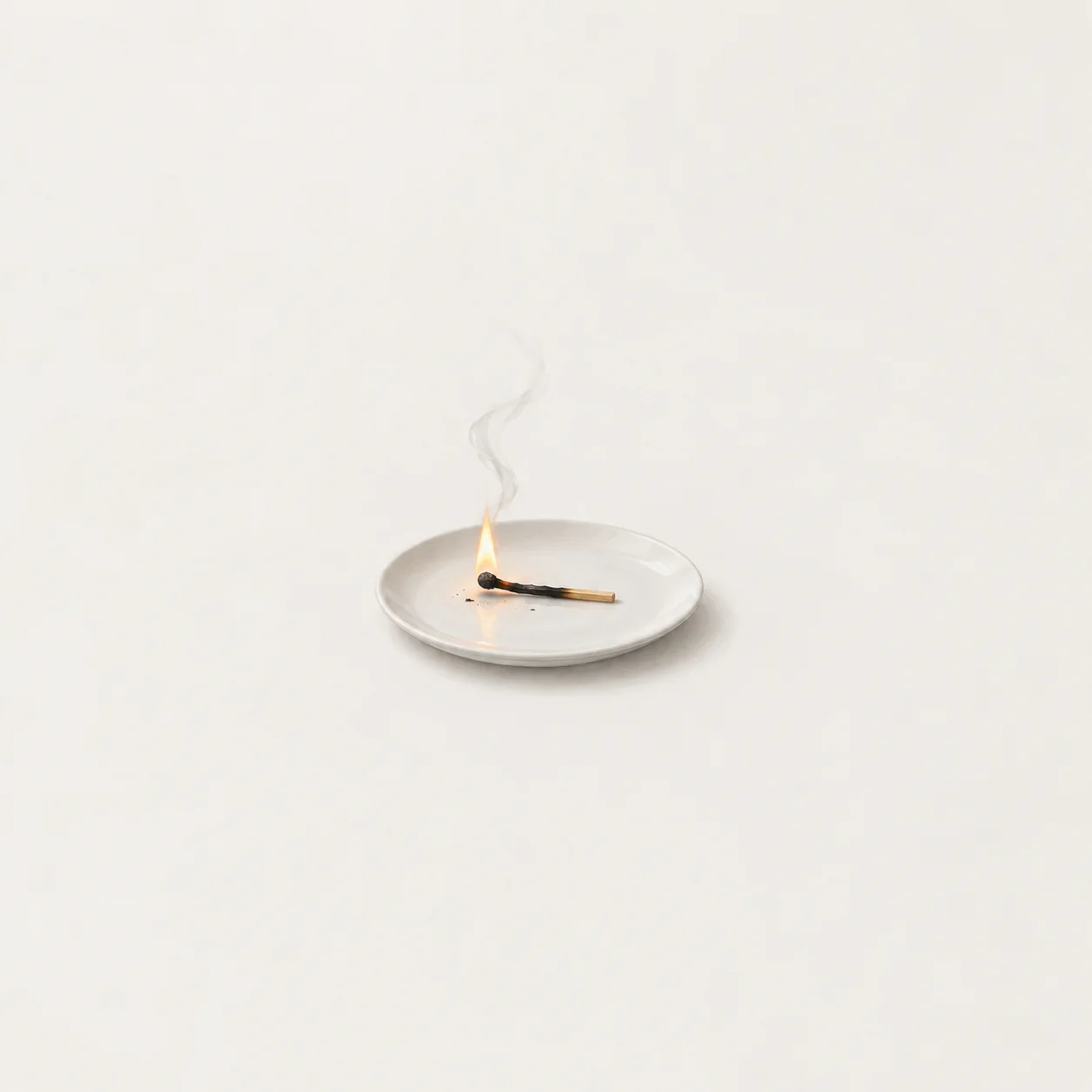 A partly burned match lies alight on a white plate, with a thin wisp of smoke above it.
