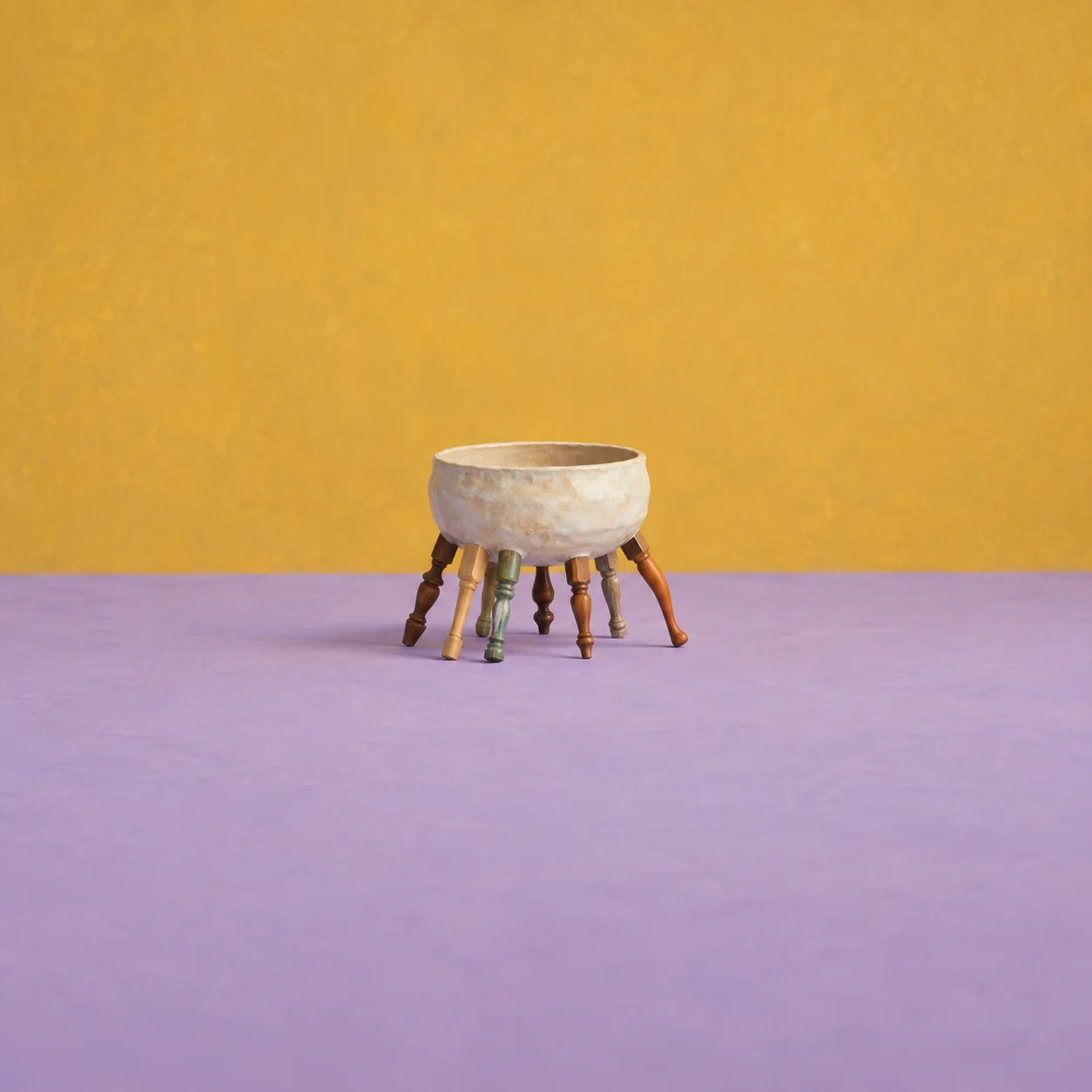 A hollow ceramic bowl stands on mismatched wooden chair legs on a lavender floor against mustard.