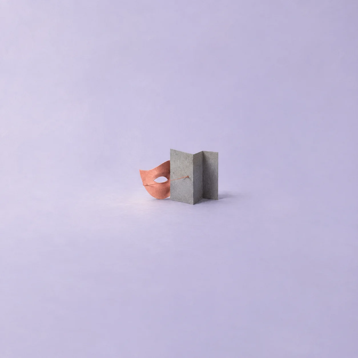 A coral paper mask peeking from behind a folded gray card screen, connected by a taut thread, on a lavender surface.