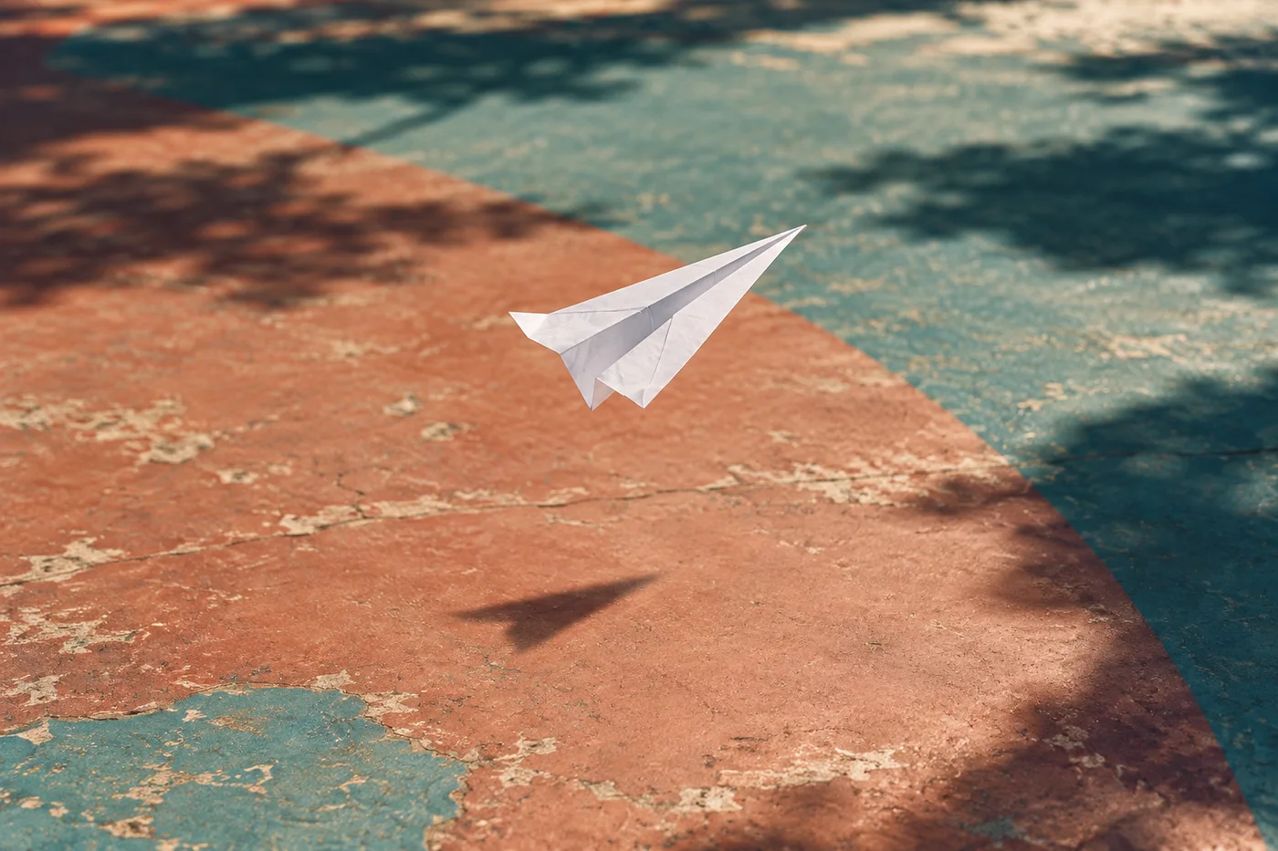 A white paper aeroplane glides above sunlit ground painted in teal and terracotta.
