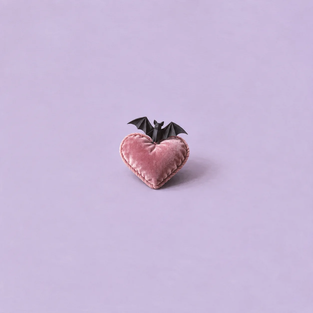 A small black paper bat perches on a pink heart-shaped cushion against pale lilac.
