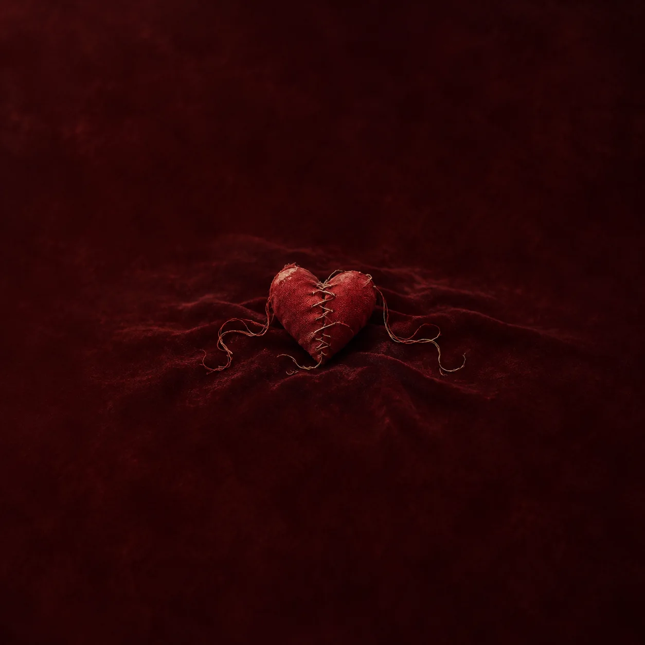 A red fabric heart with a stitched central seam and loose trailing threads rests on deep red velvet.