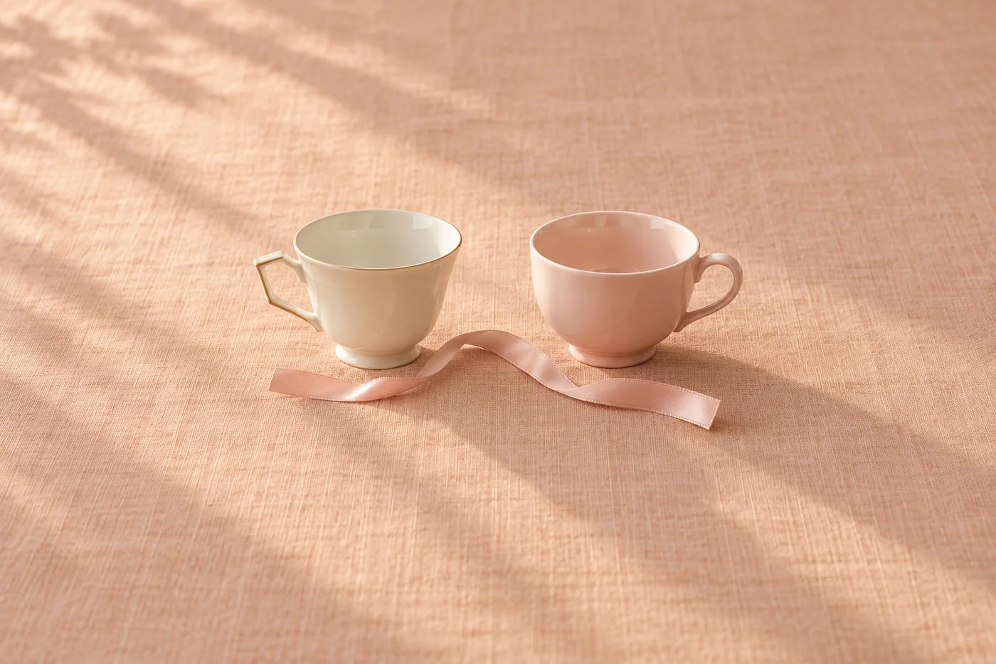 Two subtly mismatched porcelain cups and a loose pink ribbon on peach linen.