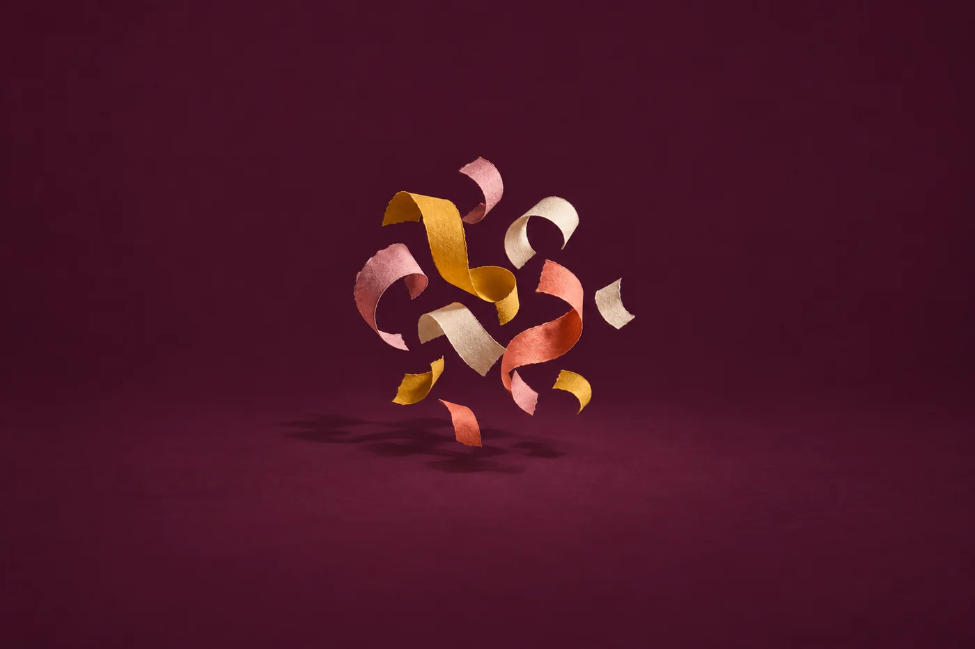 Curled strips of yellow, coral and cream paper float against a deep plum background.