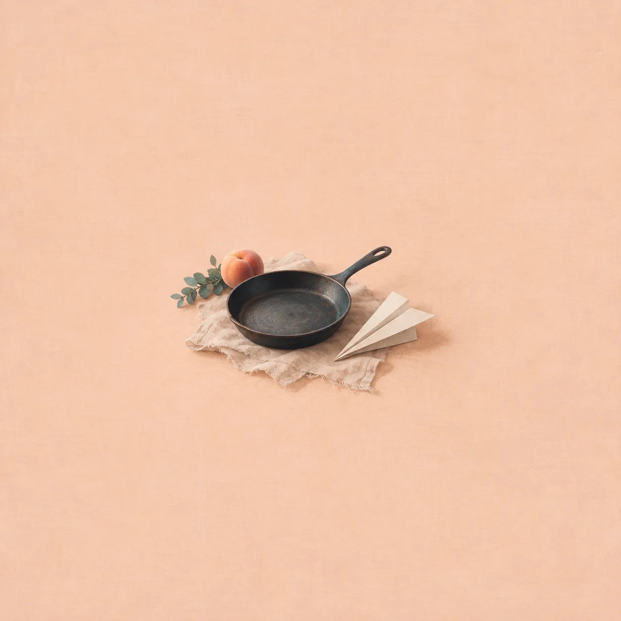 An empty frying pan beside a paper plane and a peach on rumpled cream linen against a peach background.