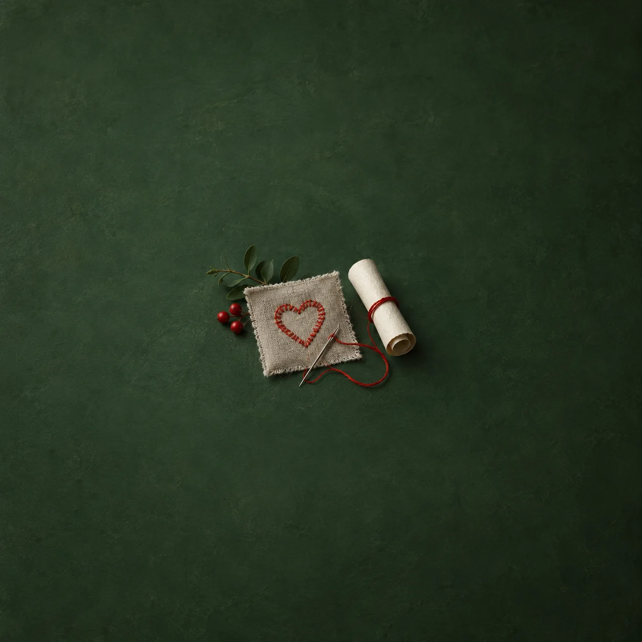 A red stitched heart and needle rest on a linen square beside a tied blank scroll and a leafy sprig.