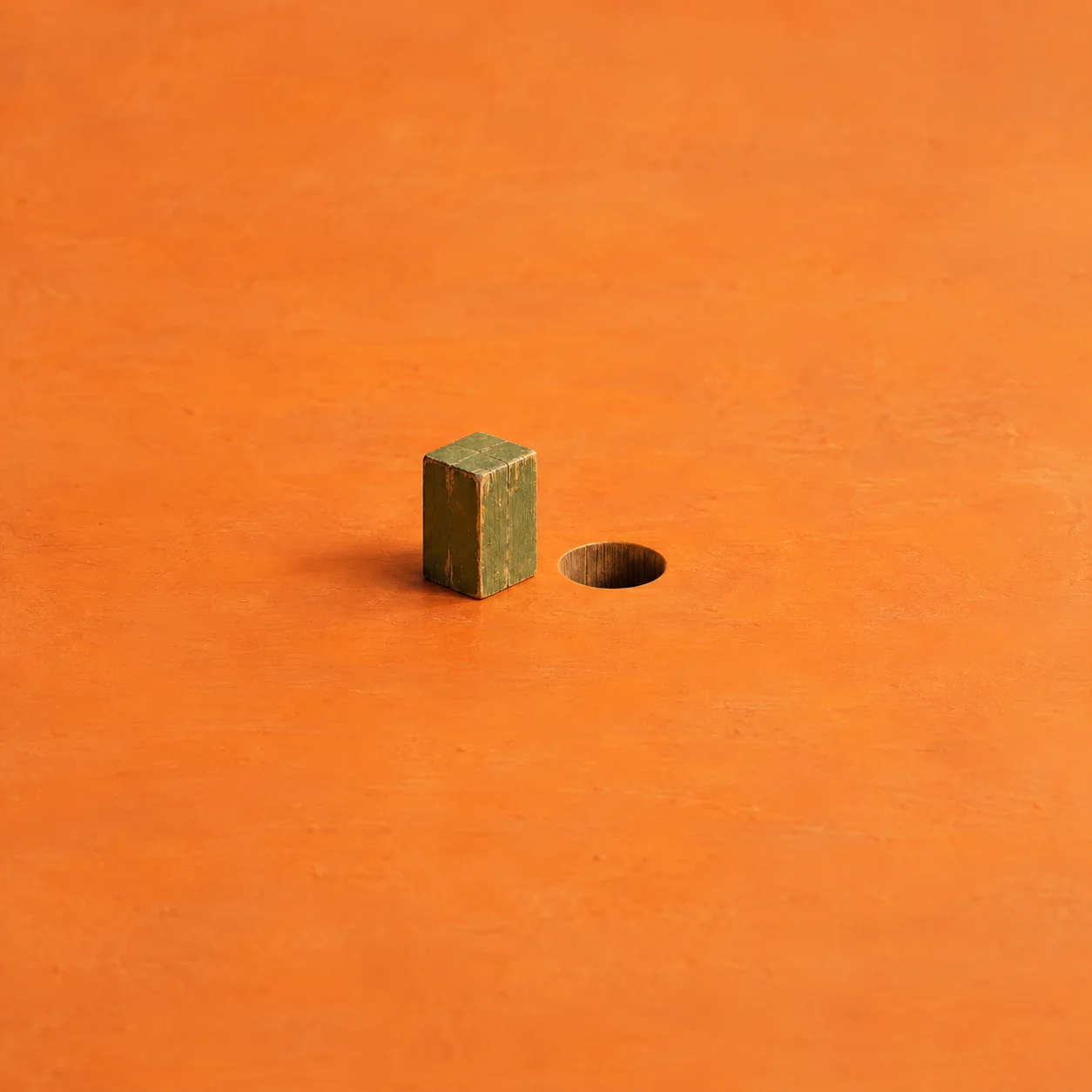A square olive-green wooden peg beside a round hole in a bright orange surface.