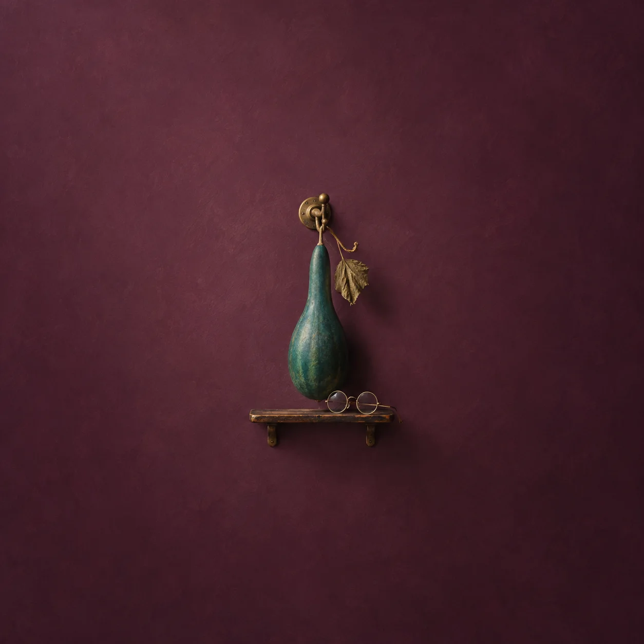 A long teal gourd hangs from a brass hook above small round spectacles on a shelf against plum.