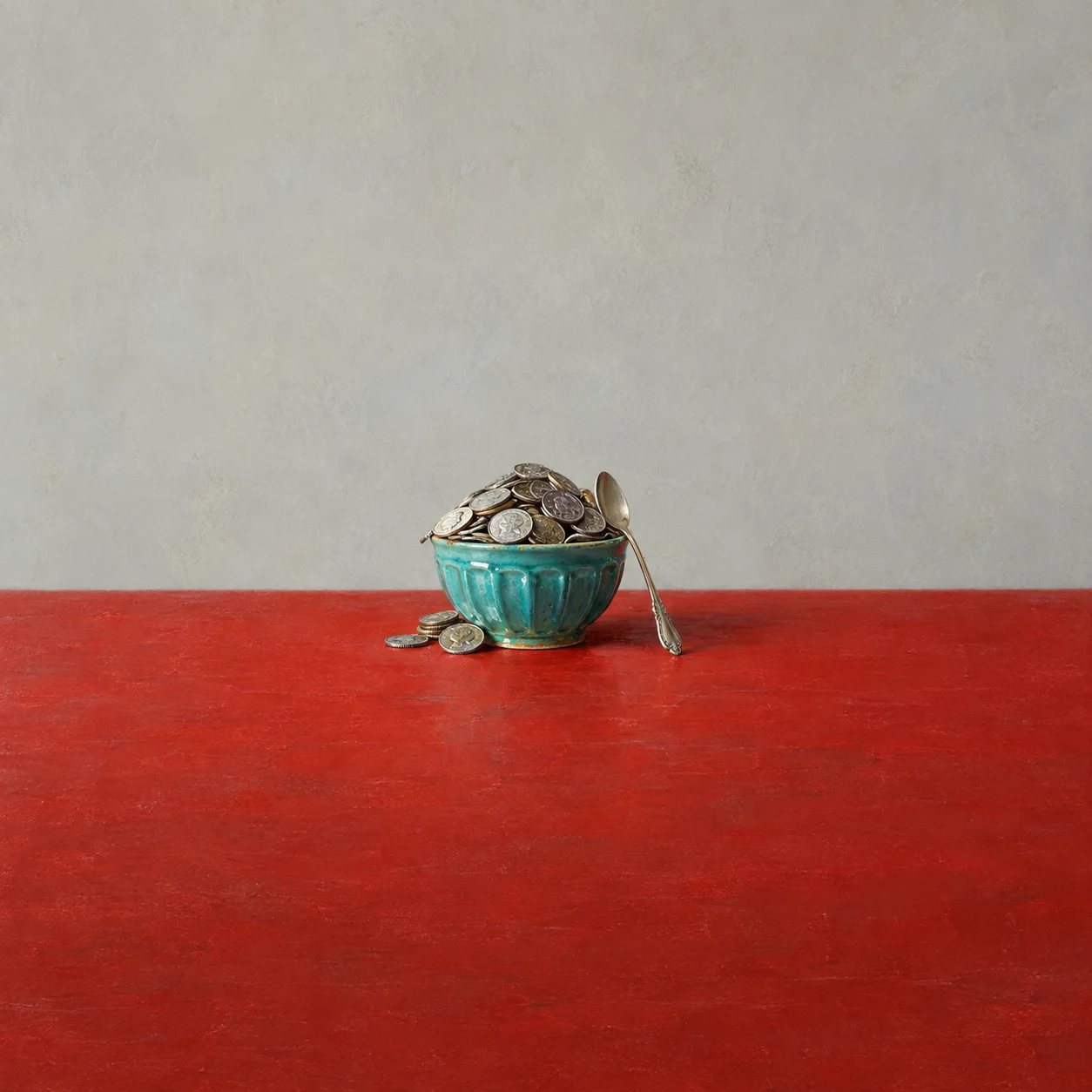 A turquoise pudding basin overflowing with silver coins with a spoon on its rim, on a red table.