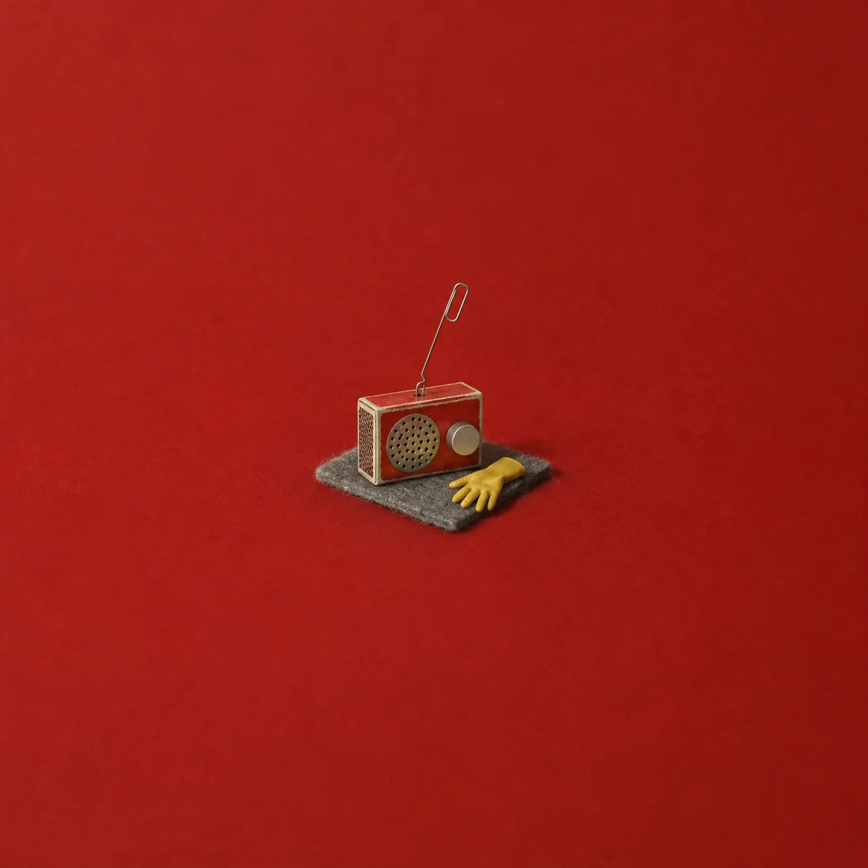 A tiny red radio with a paper-clip antenna sits beside a yellow glove on grey felt against red.