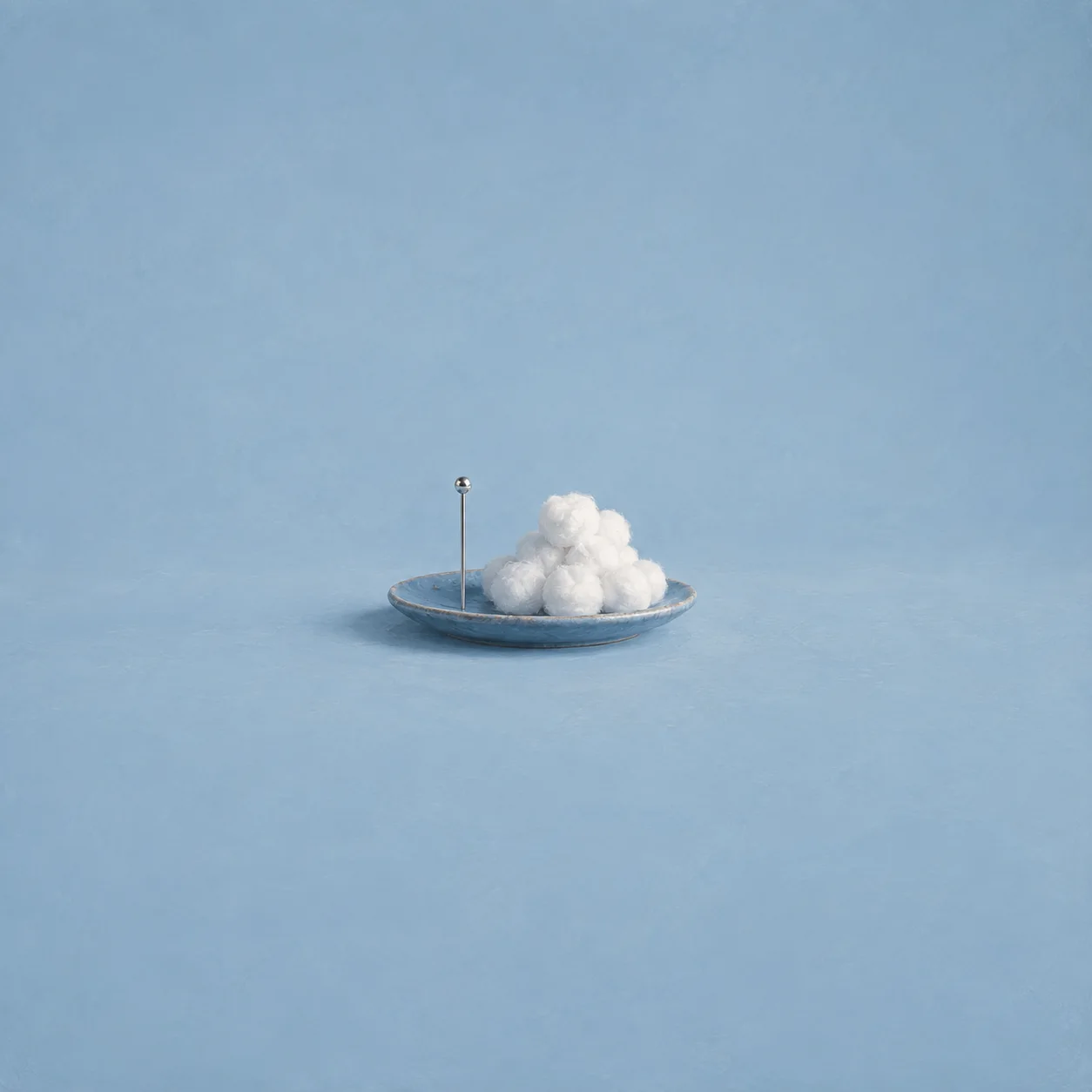A single steel pin standing upright beside a heap of cotton puffs on a blue plate.