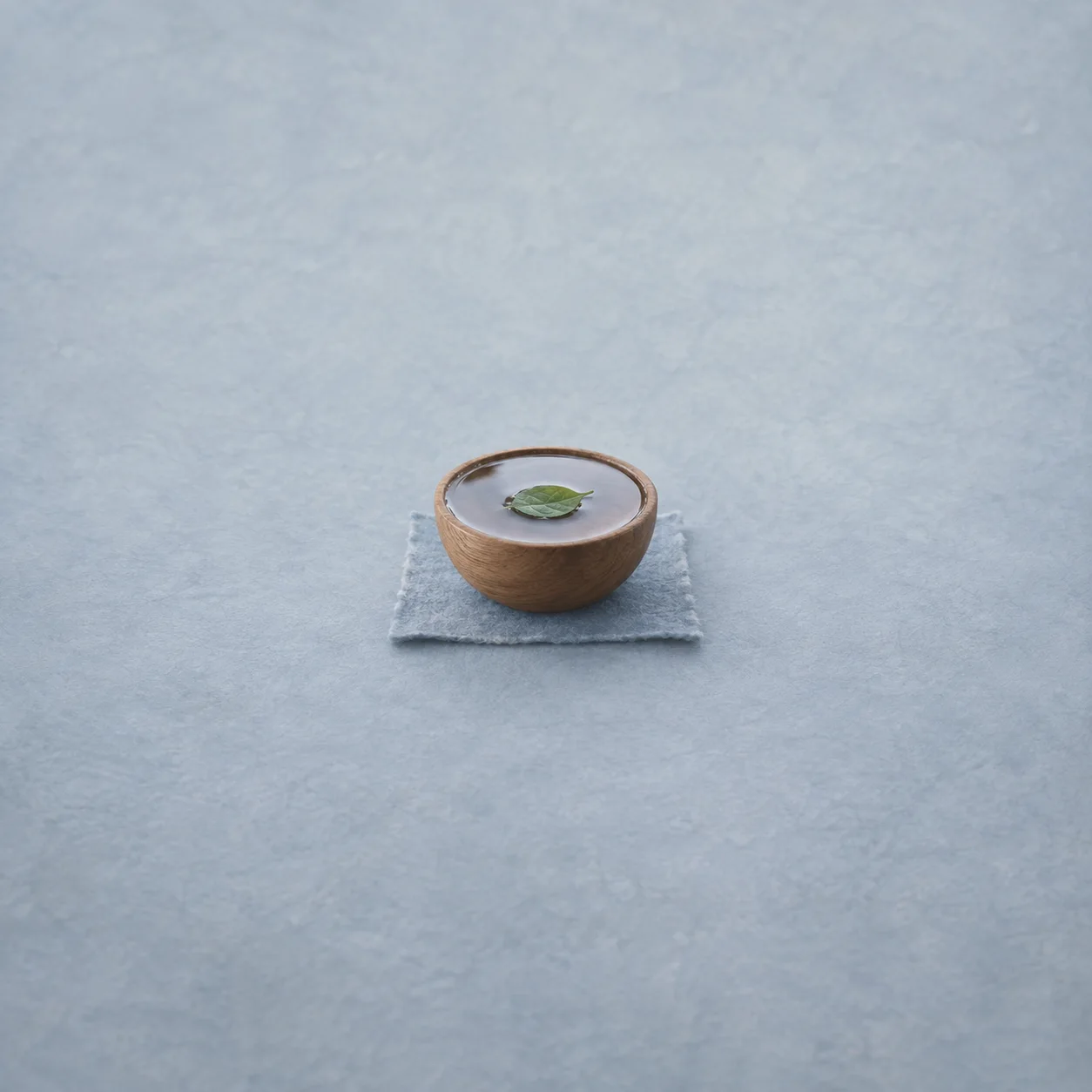 A green leaf floats in a wooden bowl of water resting on a folded blue-grey cloth.