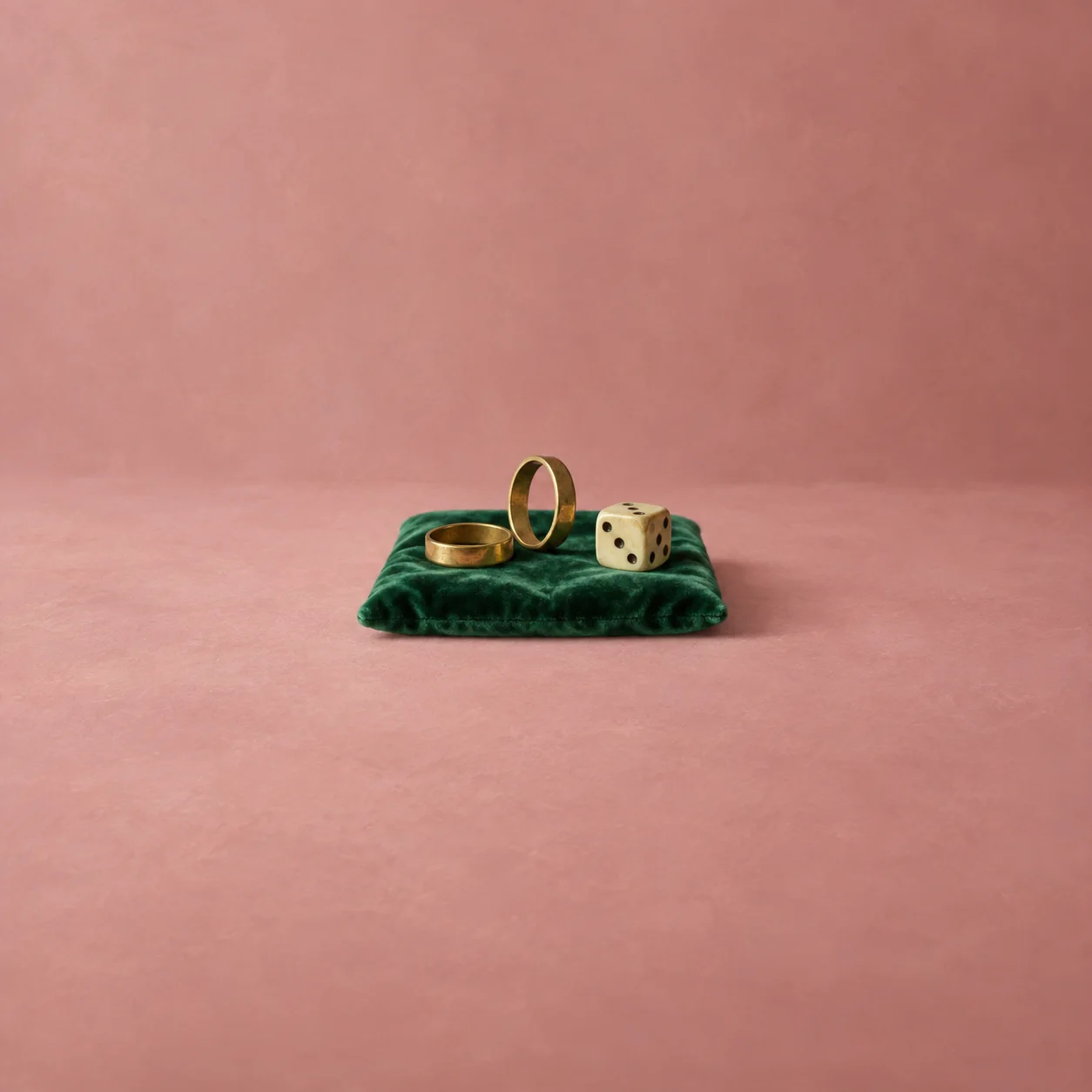 Two brass rings and an ivory die rest on folded emerald velvet against dusty pink.