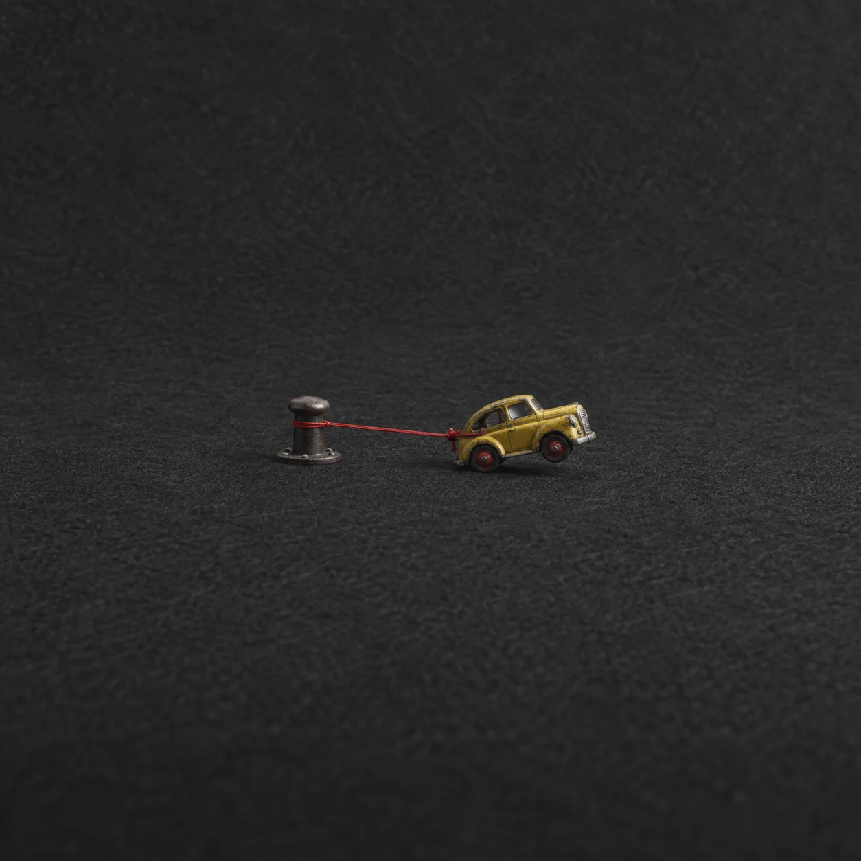 A yellow toy car is tethered to a short metal post by red string on dark grey felt.