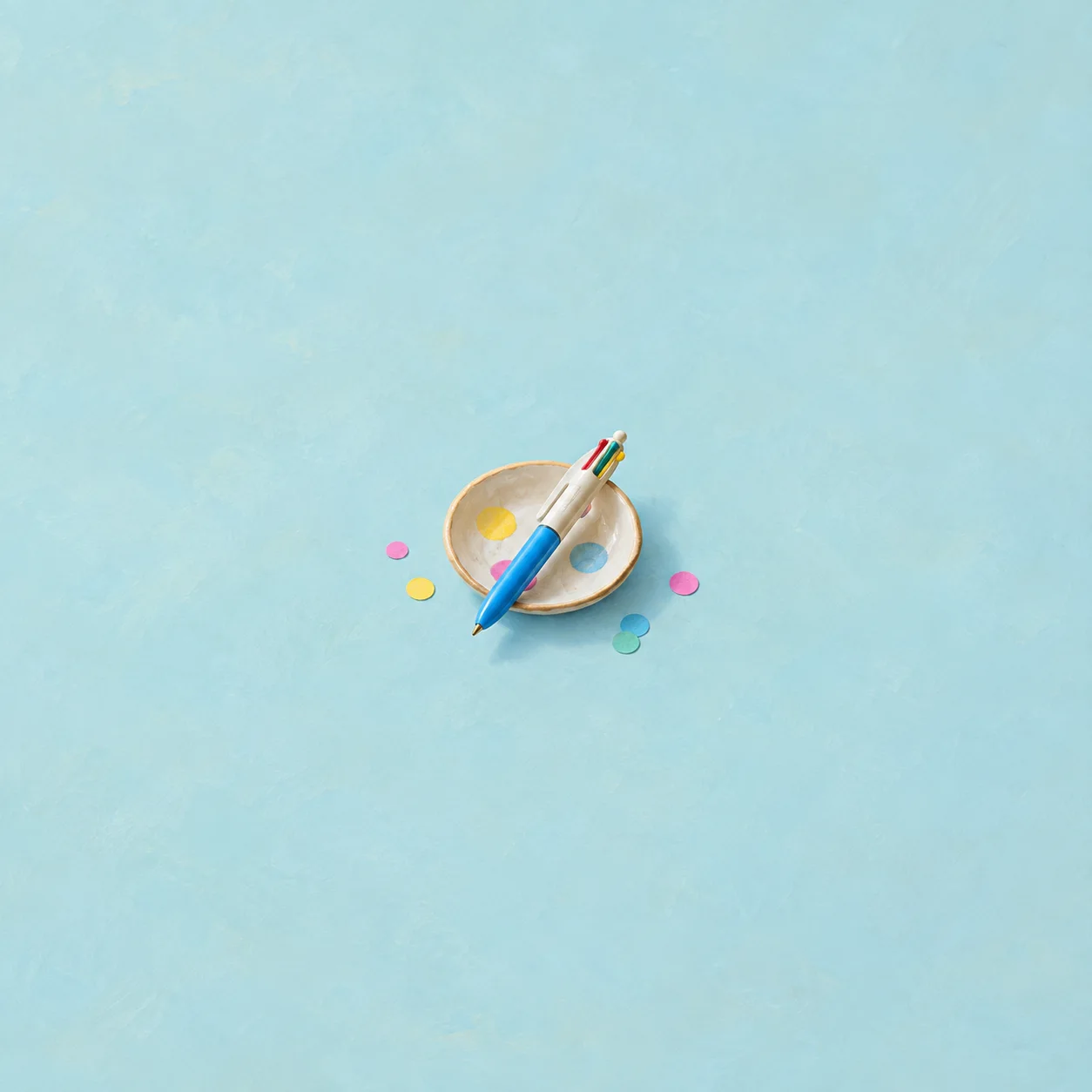 A multicolored pen rests diagonally across a shallow cream dish surrounded by bright confetti on a pale blue surface.