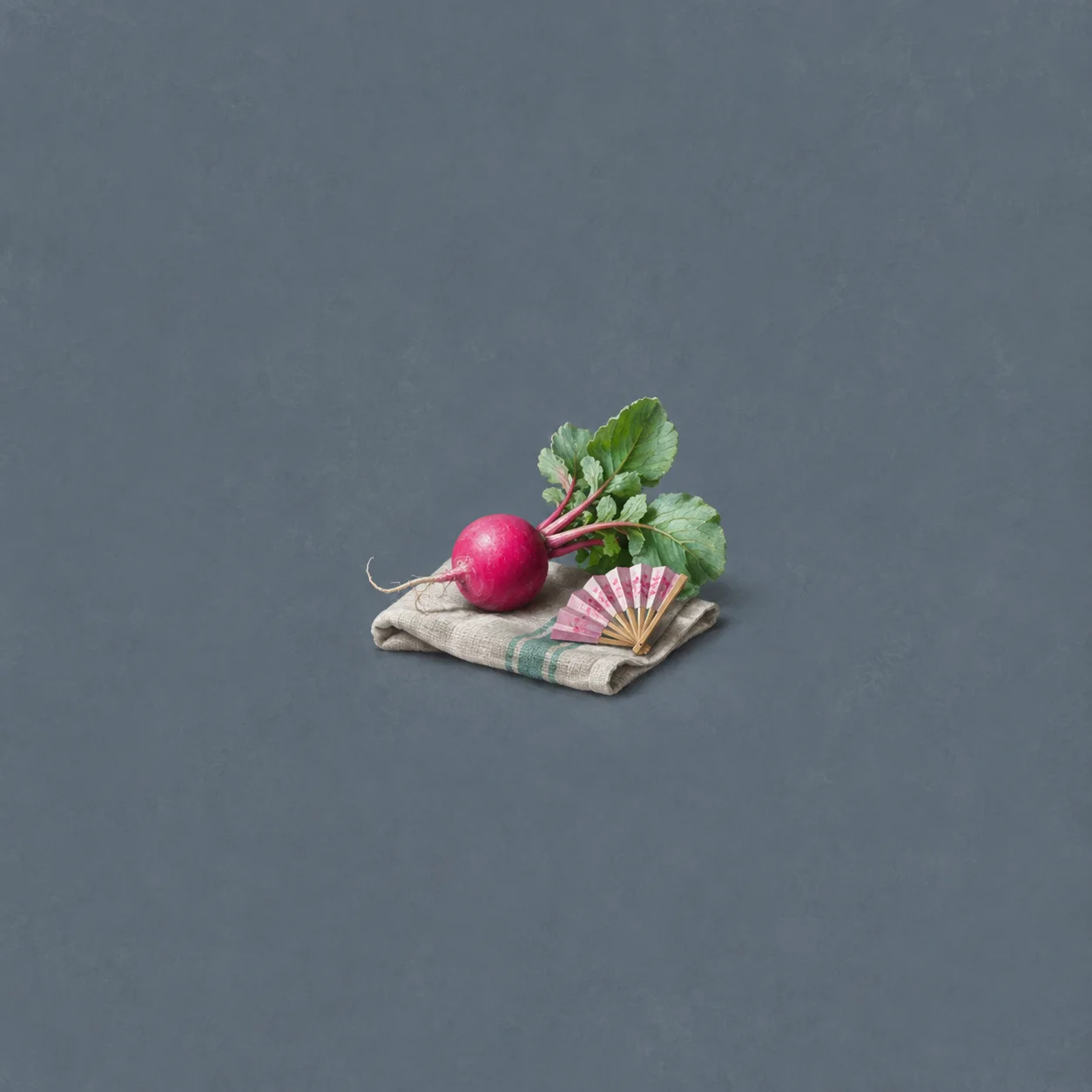 A bright pink radish with green leaves rests beside a small pink paper fan on a folded cloth.