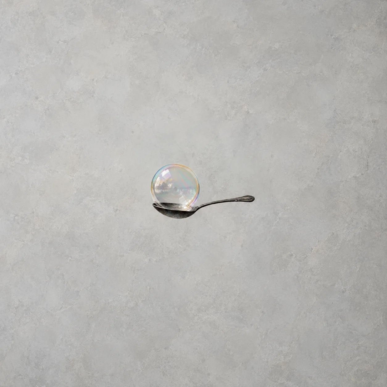 An iridescent bubble rests in the bowl of a dark metal spoon against a pale-grey background.