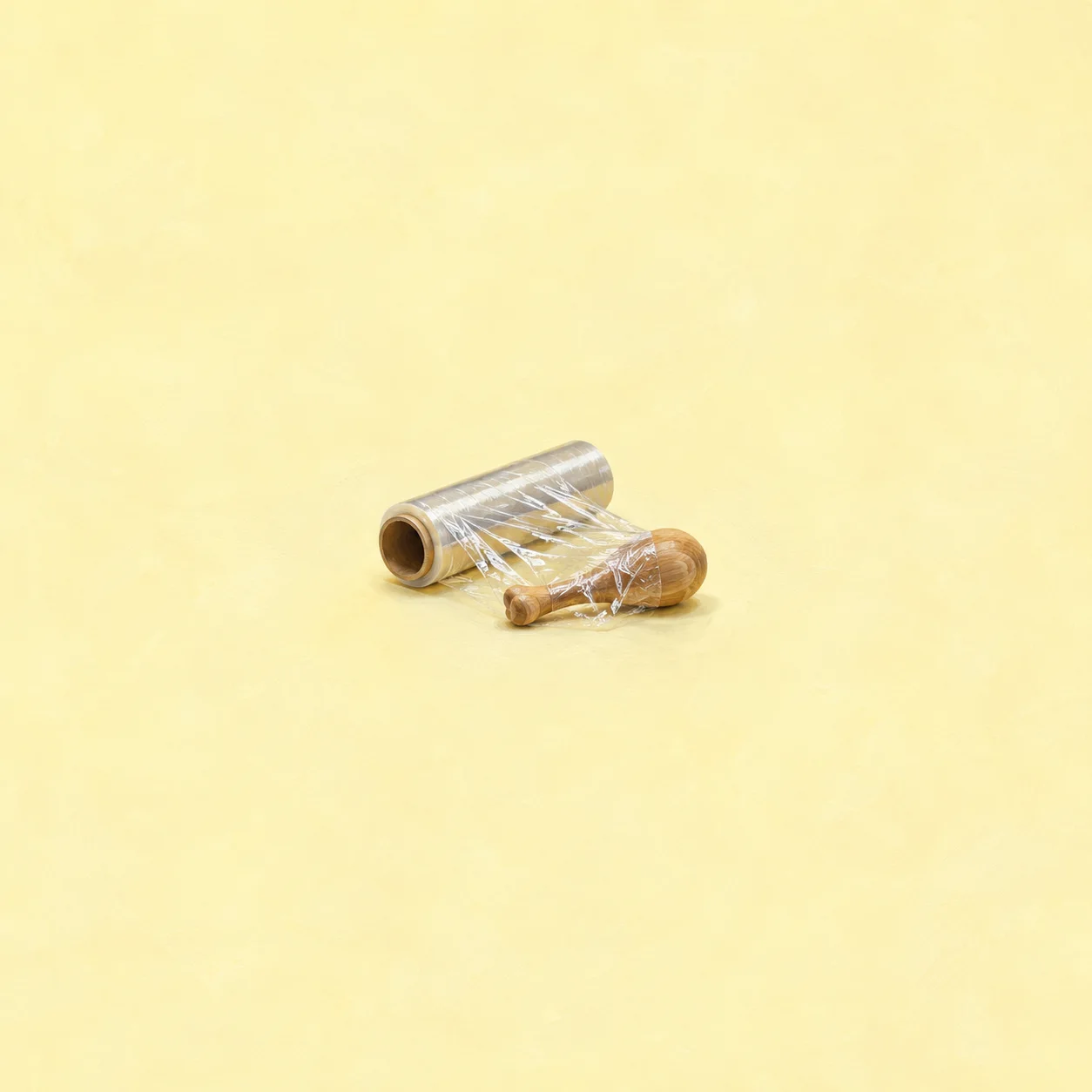 A roll of clear cling film lies partly unwrapped around a small wooden drumstick shape on a pale yellow surface.