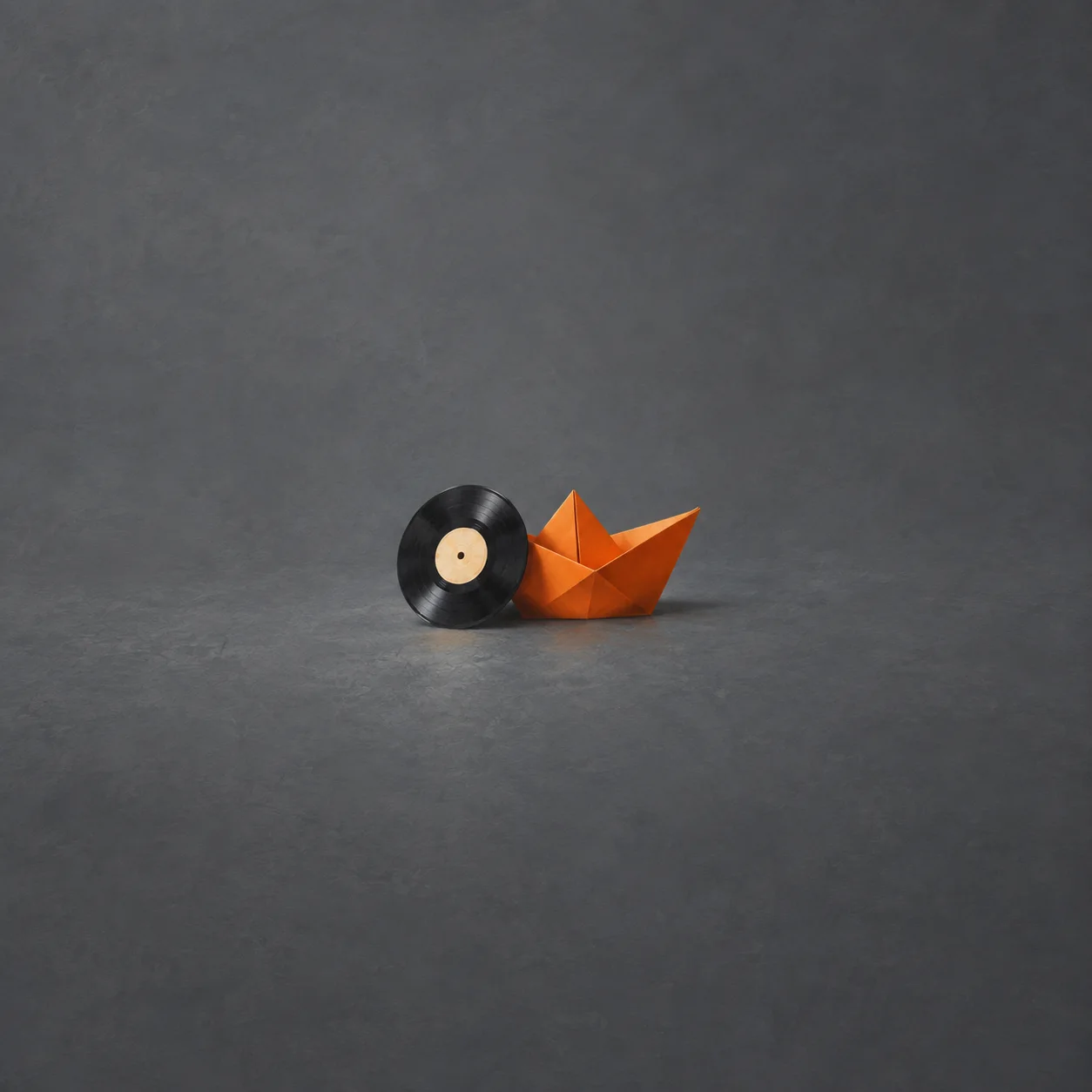 A black vinyl record leans against an orange paper boat on a slate grey surface.