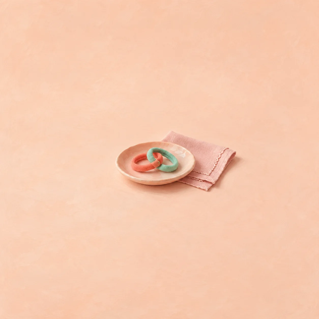 Two linked coral and mint rings on a peach saucer beside a folded pink cloth.