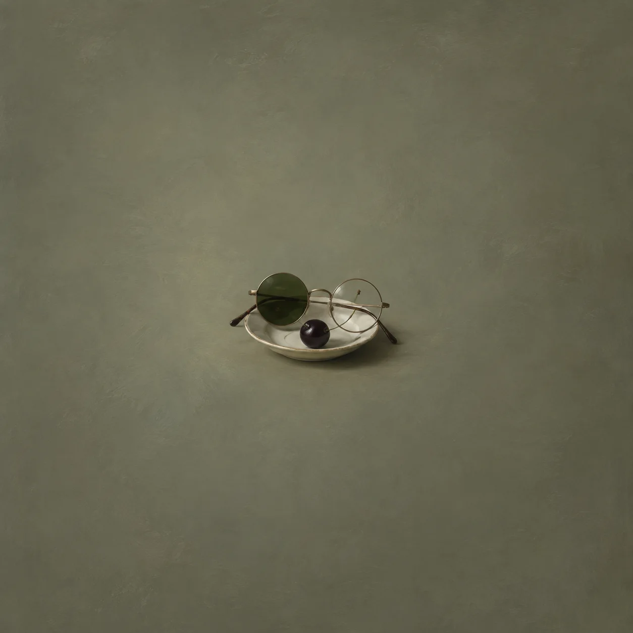 Round spectacles with one dark lens and one clear lens resting on a white saucer beside a dark cherry.