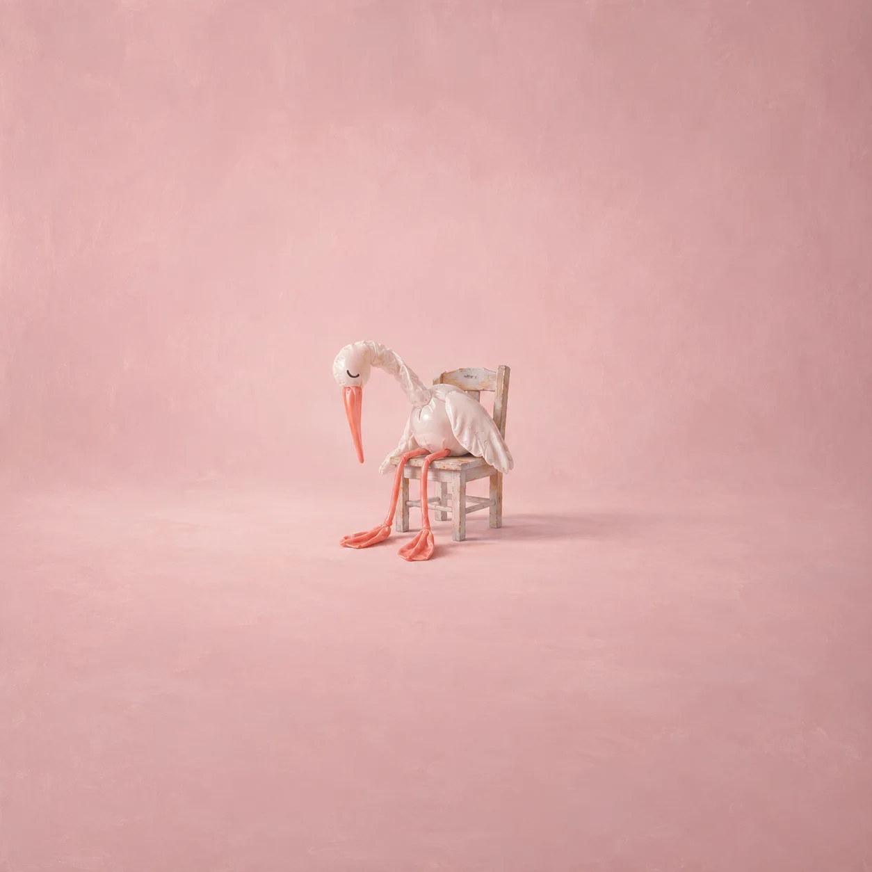 A drooping white stork-shaped balloon sits on a small chair with long orange legs against a pale pink background.