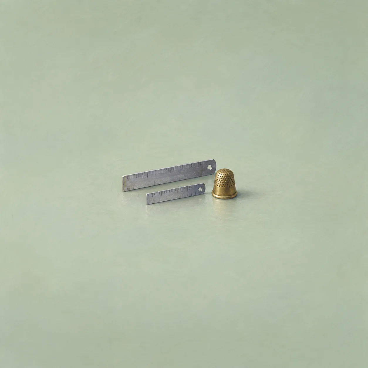 Two metal rulers of different lengths beside a brass thimble on a pale green surface.