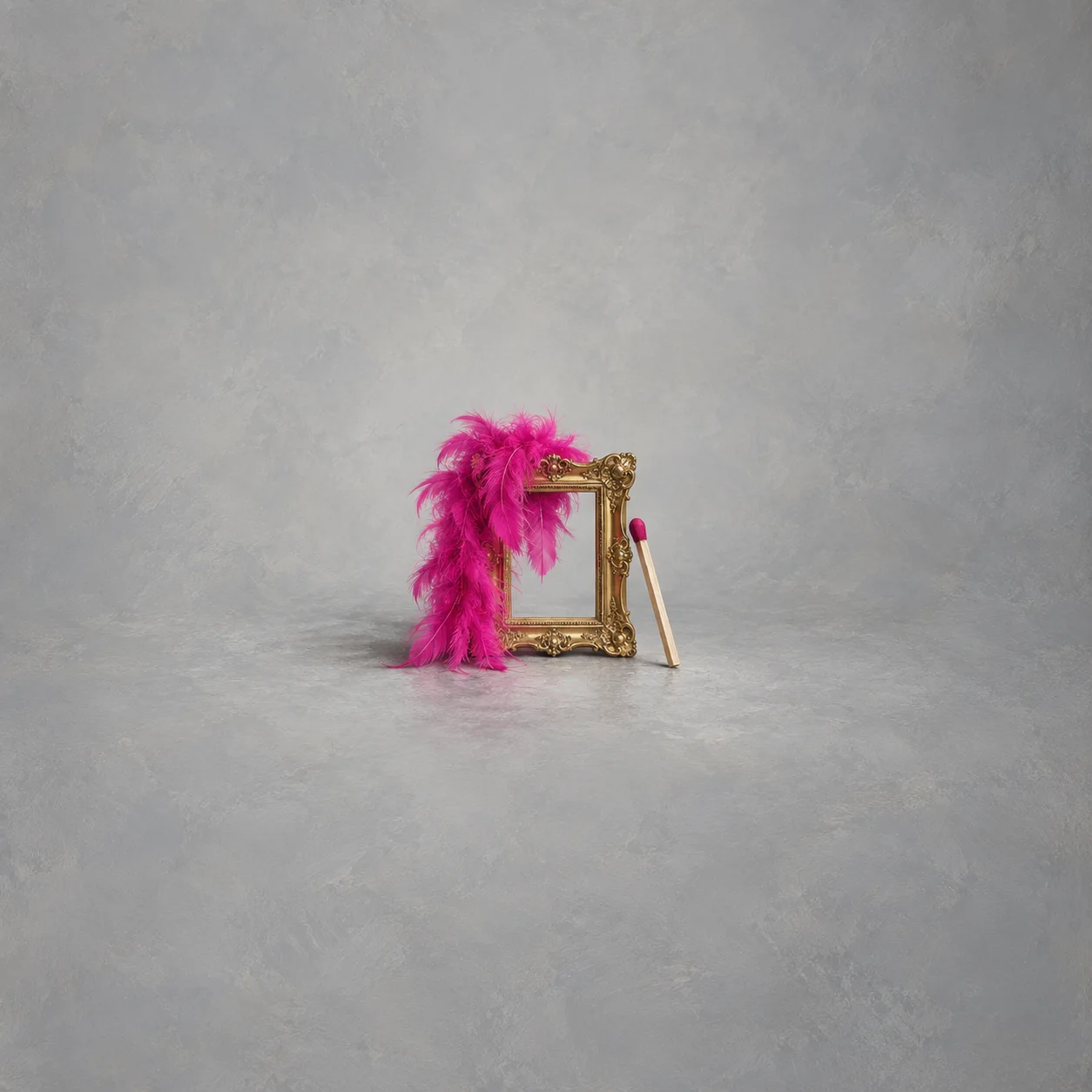A bright pink feather boa drapes over a small gold picture frame beside an unlit match against grey.