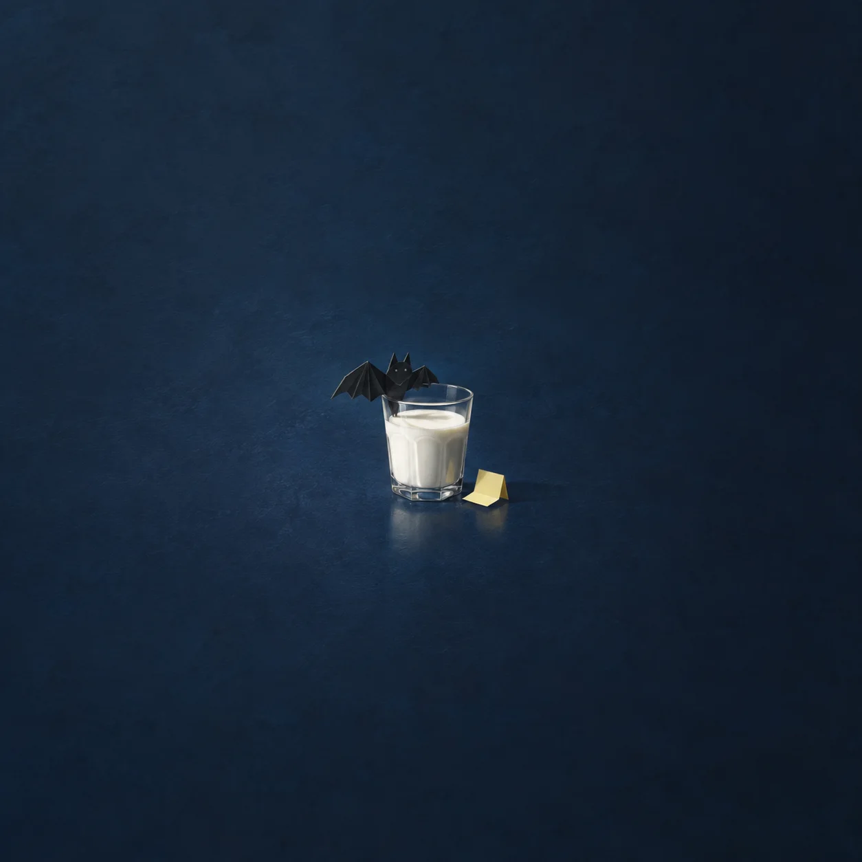 A black paper bat sitting on the rim of a glass of milk next to a folded yellow note on a midnight blue surface.