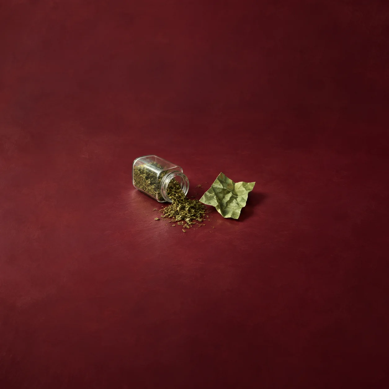 A tipped glass jar spilling dried green herbs beside a crumpled green paper on a burgundy surface.