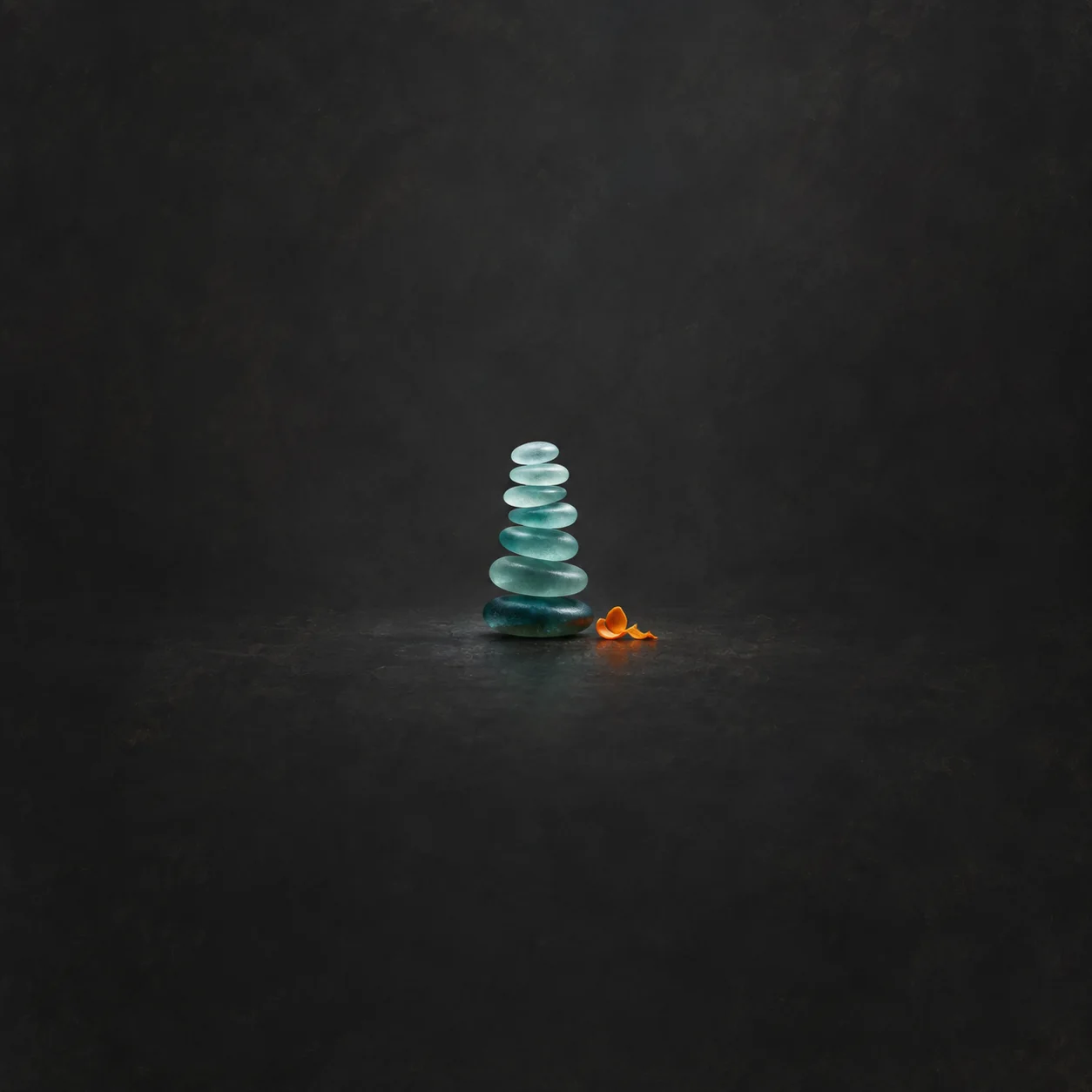 A slim stack of seven teal glass discs beside a small burst of orange confetti on a charcoal background.