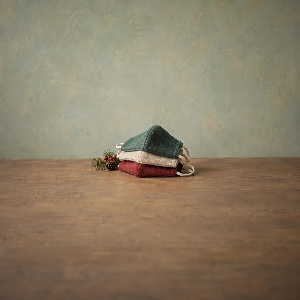Three folded cloth face coverings sit beside an evergreen sprig, berries and a pinecone.